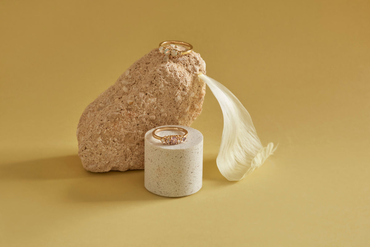 Two gold rings displayed on a textured beige stone and a small speckled white cylindrical holder, with a white feather prop, set against a solid warm yellow background.