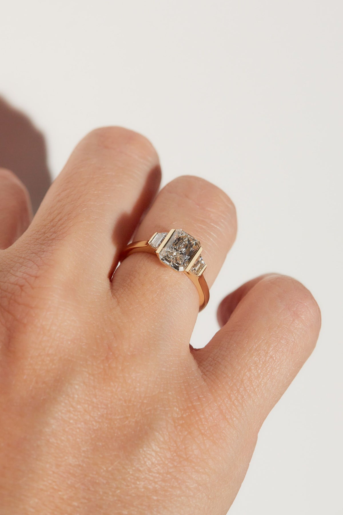 A close-up of a hand wearing a gold ring with a cushion-cut diamond on a plain light background.