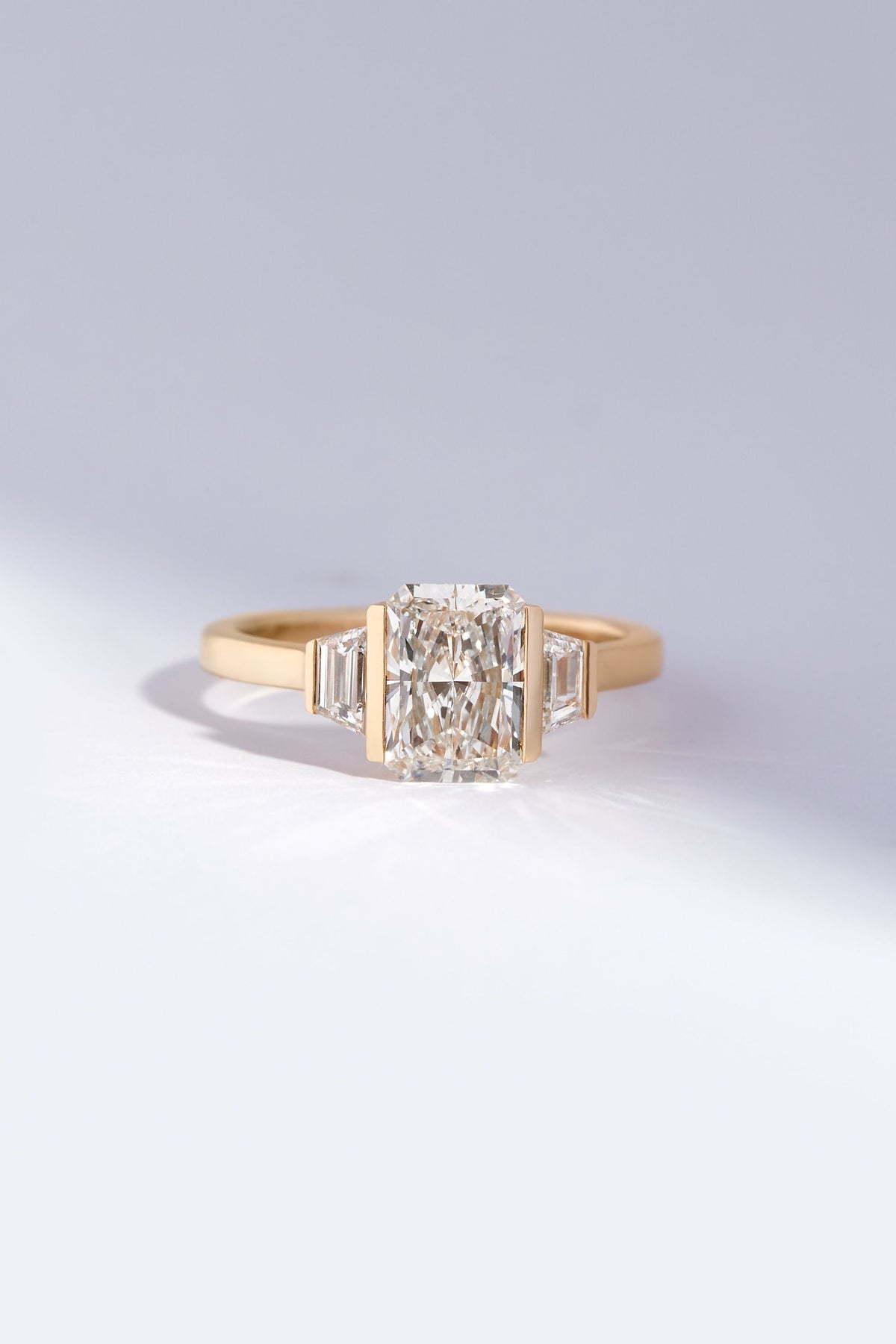 A yellow gold ring featuring a large rectangular radiant-cut center stone flanked by two baguette-cut side stones, set against a clean light gray background.