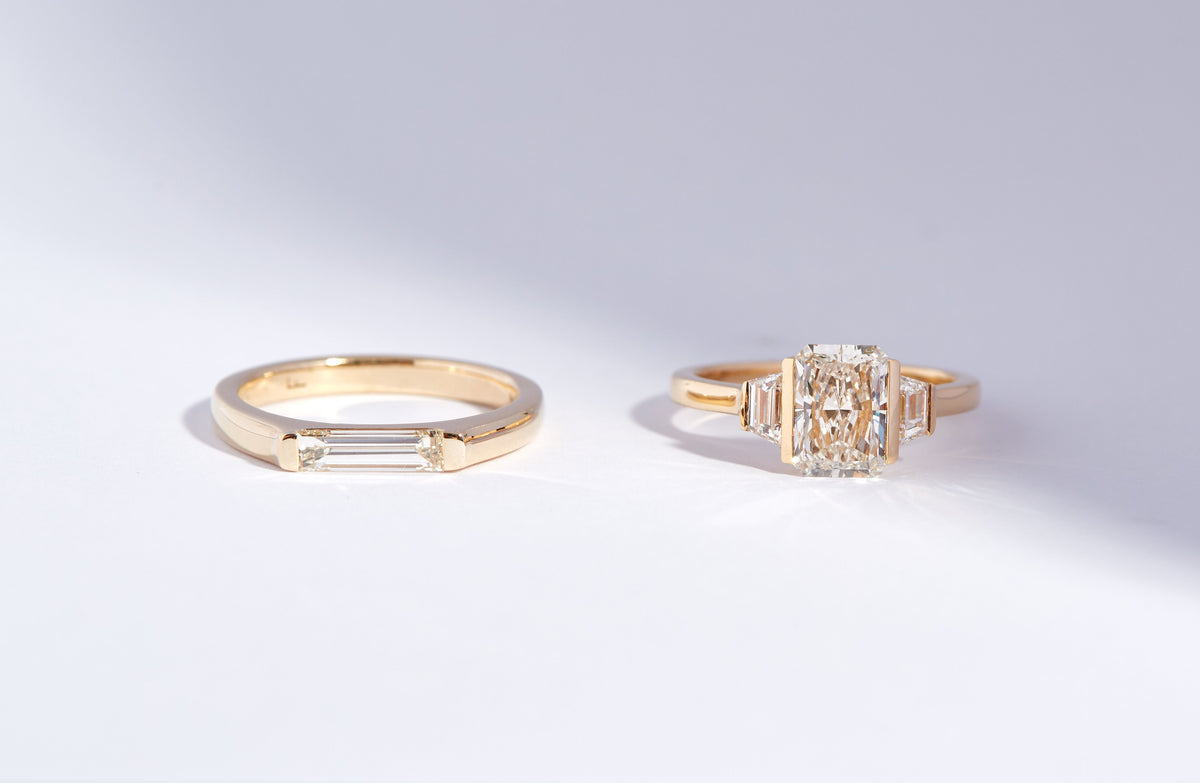 Two gold rings displayed on a clean white surface. The left ring features a smooth band with a baguette-cut diamond on one side.